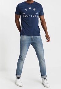 Navyblaue Baumwoll-T-Shirt mit weißem "TOMMY HILFIGER"-Logo, kombiniert mit hellblauen Jeans und weißen Sneakern, mit hochgerollten Ärmeln.