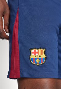 Zilās sporta šortos ar sarkanu sānu paneli, kas apakšējā labajā stūrī rotāti ar izšūtu FC Barcelona emblēmu. Strukturēta auduma.