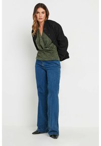 Svart texturerad bomberjacka, grön draperad topp, blå utsvängda jeans, svarta spetsiga skor. Outfiten kännetecknas av lager av tyger och en avslappnad stil.