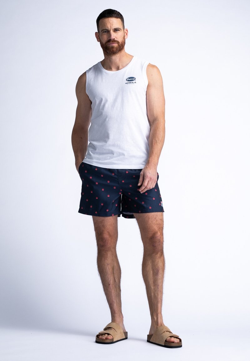 Canotta bianca in cotone senza maniche, pantaloni da nuoto blu navy con motivi floreali rossi e sandali color beige. Design semplice, vestibilità casual e aspetto versatile.