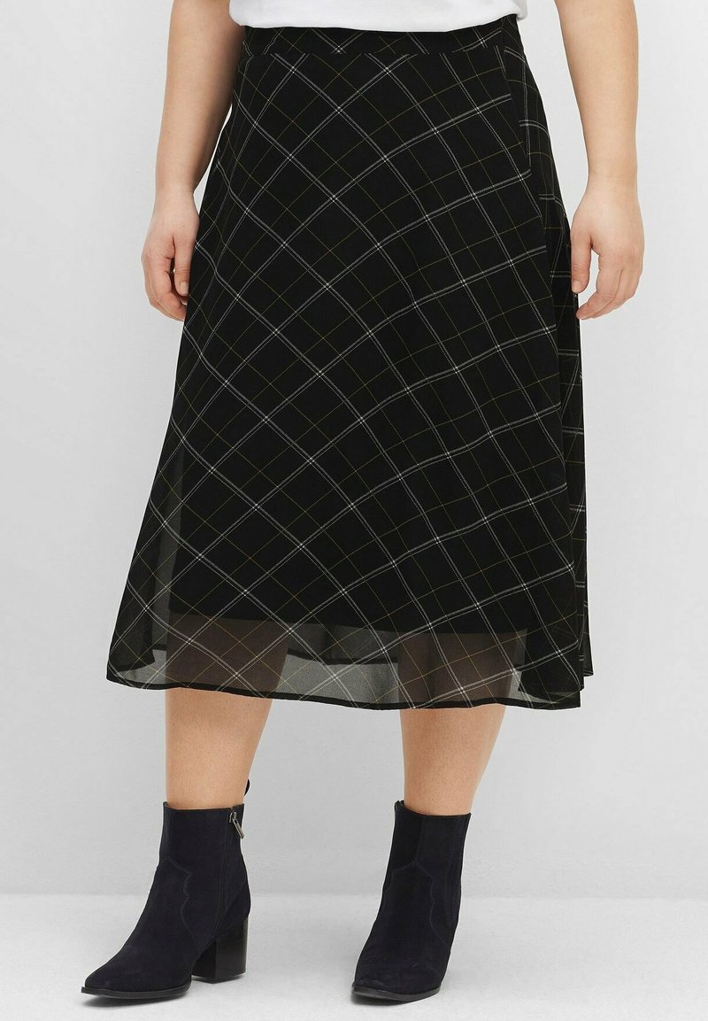 Jupe midi noire avec des lignes à carreaux blanches portée avec des bottines noires et un haut blanc, vue sur une personne debout de la taille vers le bas.