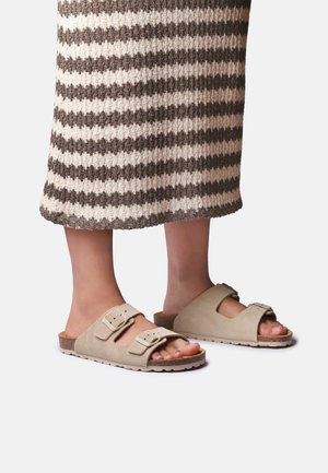Pieds chaussés de sandales slides beige à double bride avec semelles en liège, portées avec une longue jupe tricotée présentant des rayures horizontales marron et crème.