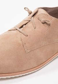 Chaussure lacée en suède beige à la texture lisse, avec des coutures ton sur ton et un bout arrondi, montée sur une semelle en caoutchouc de couleur claire.