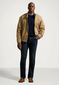 Tan bomberjakke over en marineblå kabelstrikket genser, kombinert med mørke jeans og brune loafers. Merkbare detaljer inkluderer glidelås og ribbestrikkede mansjetter.