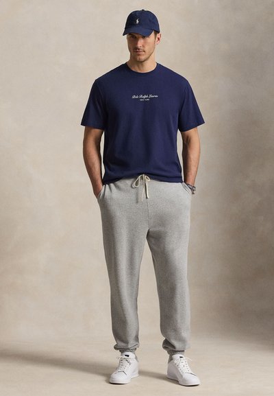 Άνδρας που φοράει σκούφο και μπλουζάκι Polo Ralph Lauren σε σκοτεινό μπλε, ανοιχτό γκρι φόρμα, λευκά αθλητικά παπούτσια, στέκεται με τα χέρια στις τσέπες.