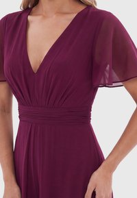 Femme portant une robe violet profond avec un décolleté en V, un détail froncé à la taille et des manches courtes transparentes et flottantes.