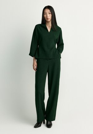 Woman wearing a dark green long-sleeve top and matching wide-leg pants, standing with one hand in her pocket and black heeled shoes.