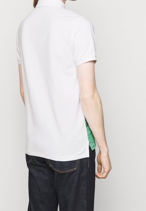 Osoba ubrana w gładką białą koszulkę polo z krótkim rękawem i ciemne dżinsy, widziana od tyłu na jednolitym jasnym tle.