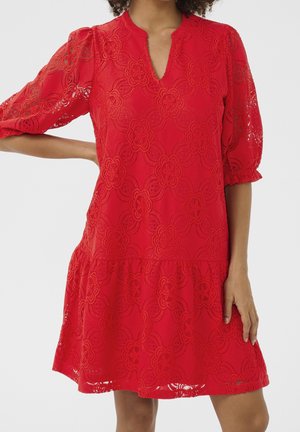 Czerwona sukienka z kwiatowym koronkowym wzorem, bufkami do łokcia i małym dekoltem w kształcie litery V, noszona przez osobę z jedną ręką na biodrze.