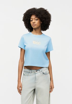 Νεαρή γυναίκα με σγουρά μαλλιά που φοράει ανοιχτό μπλε κοντομάνικη μπλούζα με στάμπα «1986» και ανοιχτόχρωμα τζιν ψηλόμεσα, σε λευκό φόντο.