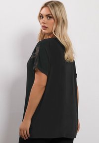 Top nero realizzato in morbido tessuto, con maniche corte, dettagli in pizzo sulle spalle e un design sfiancato, visibile dal retro.