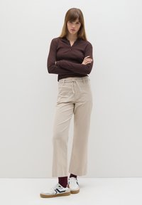 Bruine lange mouwen henley-shirt, beige wijde broek met trekkoord in de taille, witte sneakers met marineblauwe accenten, staande tegen een witte achtergrond.