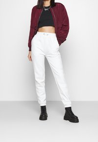 Kvinna iklädd en kort svart Nike-top, marinblå bomberjacka, vita sweatpants och svarta platåkängor som står mot en vit bakgrund.