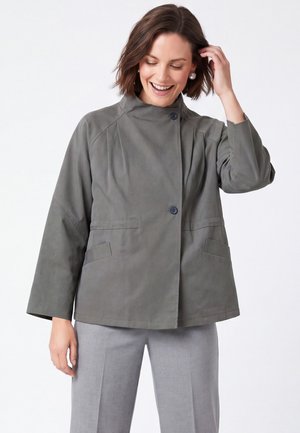 Donna sorridente, indossa una giacca grigia ampia con bottoni, tasche e maniche larghe, abbinata a pantaloni grigio chiaro su sfondo bianco.