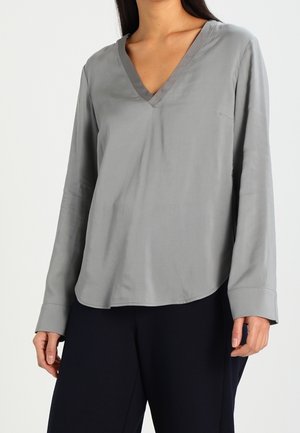 Kvinde iført en lysegrå V-hals bluse med lange ærmer, kombineret med løstsiddende sorte bukser mod en hvid baggrund.