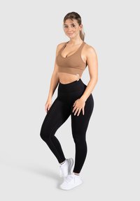 Brune geribbelde sportbeha in combinatie met hoog aansluitende zwarte leggings en witte sportschoenen. Model poseert tegen een grijze achtergrond.