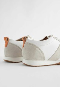 Wildleder und synthetische weiße Sneakers mit beigem Absatz, ausgestattet mit orangefarbenen Akzenten und einer schlaufenförmigen Zuglasche auf der Rückseite für einfaches Anziehen.