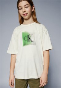 Krämfärgad t-shirt med korta ärmar, prydd med ett grönt och svart grafiskt tryck på framsidan. Tillverkad av mjukt bomullsmaterial.
