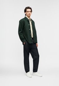 Πράσινο σακάκι με κουμπιά, ανοιχτό κίτρινο πουκάμισο από κάτω, μαύρο παντελόνι και λευκά αθλητικά παπούτσια. Λείες υφές, casual εφαρμογή, καθαρές γραμμές.