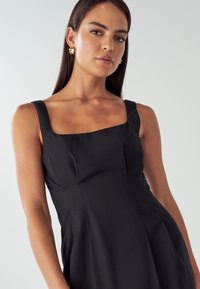 Črna, oprijeta obleka s kvadratnim izrezom in širokimi naramnicami. Gladka tekstura z osnovno bodico. Zlate obroče kot dodatek.