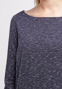 Mörkgrå långärmad topp med bred u-ringning, med ett texturerat tyg som har ljusa fläckar. Mjuk, avslappnad passform med minimal sömnad.