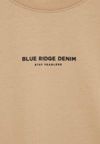 Tissu tricoté beige avec texte noir "BLUE RIDGE DENIM" au-dessus d'un texte plus petit "STAY FEARLESS" centré sous l'encolure.