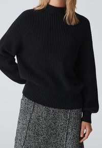 Zwarte geribbelde gebreide trui met een hoge halslijn en lange mouwen, gecombineerd met een gestreepte grijze rok met een visgraatpatroon.