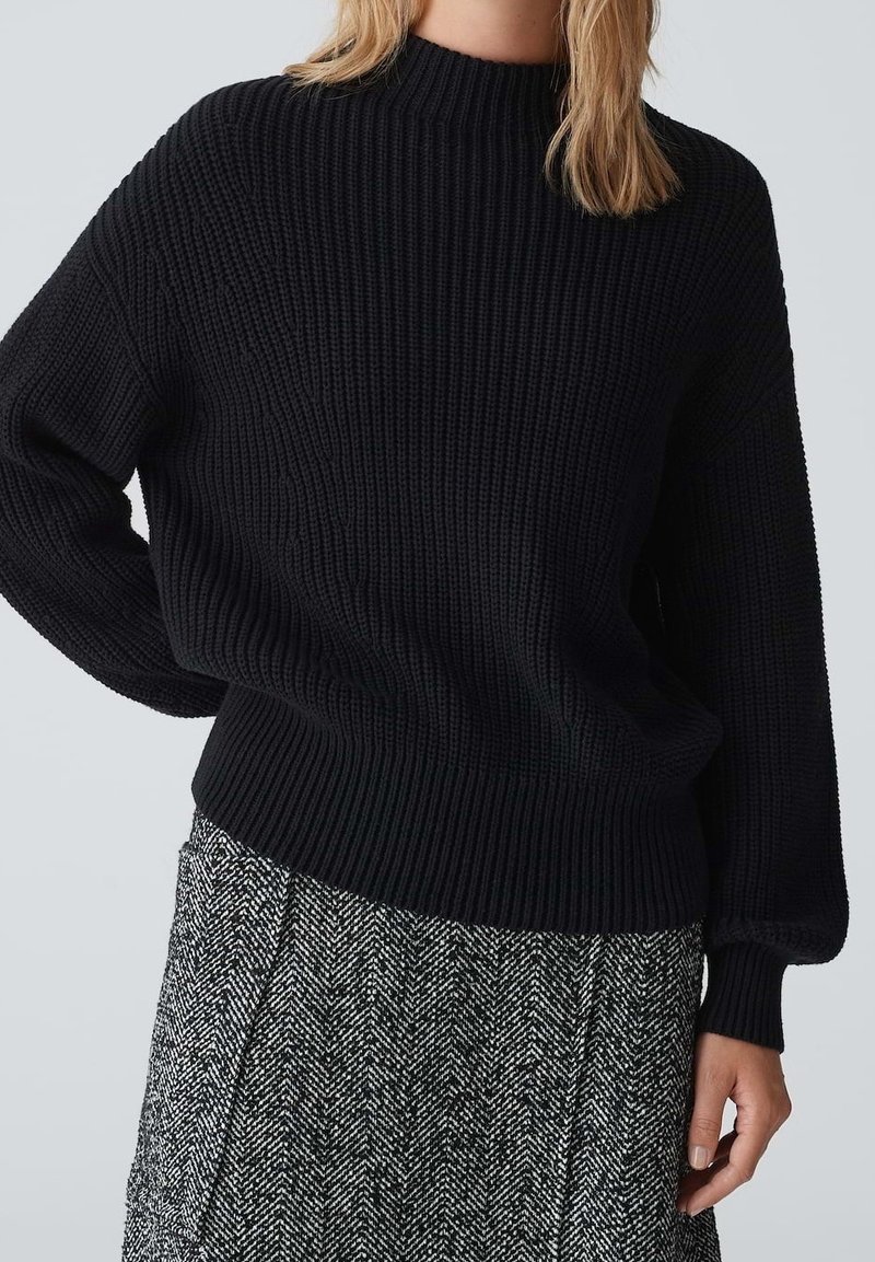 Zwarte geribbelde gebreide trui met een hoge halslijn en lange mouwen, gecombineerd met een gestreepte grijze rok met een visgraatpatroon.