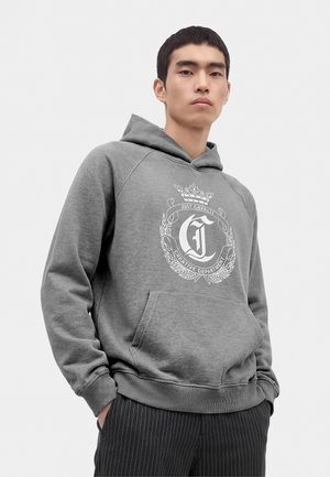 Joven con sudadera gris con capucha, con texto gráfico blanco y diseño, de pie con las manos en los bolsillos frente a un fondo liso.