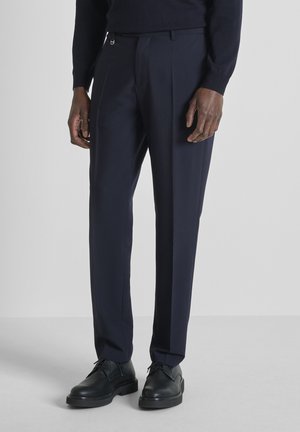 Donkerblauwe, op maat gemaakte broek gedragen met zwarte leren veterlaarzen, gezien bij een staand persoon tegen een effen lichte achtergrond.