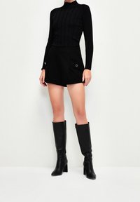 Woman wearing black ribbed long-sleeve top, high-waisted black shorts with buttoned pockets, and knee-high black leather boots standing against white wall.