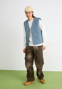 Gilet en coton bleu avec un devant ouvert et des poches latérales, porté sur une chemise blanche à manches longues et un pantalon cargo marron, associé à des bottes beiges.