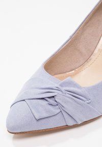 Ballerine pointue en tissu violet clair avec un nœud décoratif à l'avant. La chaussure présente une texture douce et une doublure intérieure couleur camel.
