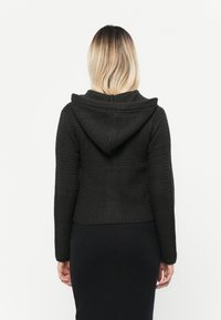 Svart strikket hettegenser med en teksturert mønster, tettsittende design og hette. Båret over et svart plagg, som viser en cropped lengde.