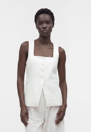 Femme à la peau foncée portant un haut blanc sans manches boutonné avec un décolleté carré et un pantalon blanc à motifs, debout devant un fond uni.