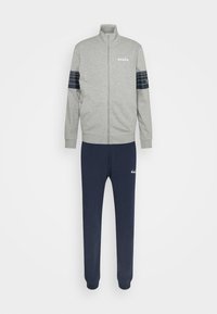 Veste de survêtement grise zippée avec des rayures horizontales bleu marine sur les manches, associée à un pantalon de survêtement bleu marine avec un petit logo blanc sur la cuisse.