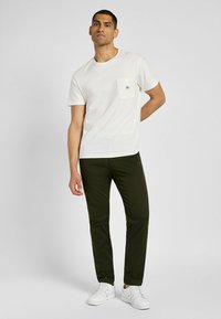 Camiseta de algodón blanca con un bolsillo en el pecho, combinada con pantalones verde oscuro y zapatillas blancas. Diseño sencillo con mangas cortas y un ajuste relajado.