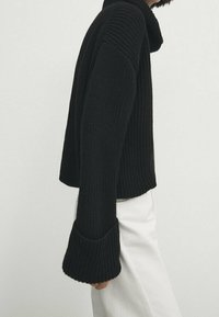Black knitted sweater with a high collar, ribbed texture, and wide cuffs. Worn with white slightly flared pants.