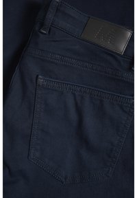 Jean en denim bleu foncé montrant la poche arrière avec des coutures détaillées et un patch en cuir noir de la marque sur la ceinture.