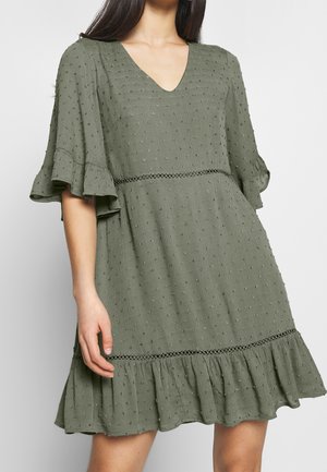 Kobieta w oliwkowej, teksturowanej sukience z dekoltem w serek, z falbanami na rękawach i falbanianym dołem, ukazująca środkową część uda i częściowo ramię.