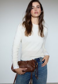 Maglione bianco a maglia con maniche lunghe, abbinato a jeans blu a vita alta. La modella tiene in mano una borsa a coulisse in pelle marrone con dettagli dorati.