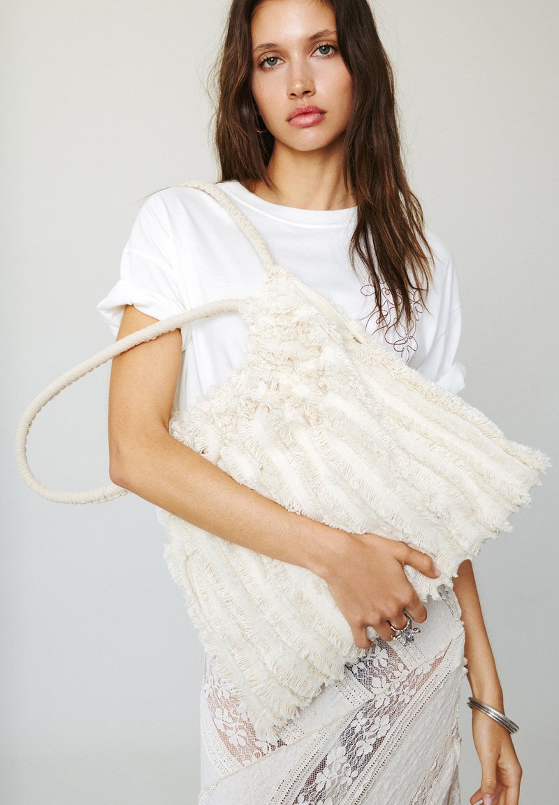 Ivory textured bag with frayed fabric stripes and soft handles, held by a person wearing a white t-shirt, featuring a lace skirt in the background.