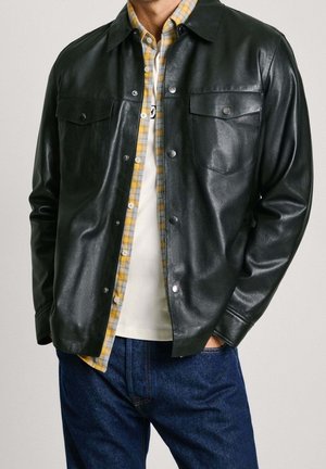 Hombre con chaqueta de cuero negra sobre camisa a cuadros amarilla y camiseta blanca, con jeans azules, manos en los bolsillos, de pie frente a un fondo neutro.