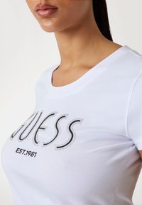 T-shirt bianco a maniche corte con un grande logo nero brillante "GUESS" e la scritta "EST. 1981" stampata sotto in nero, scollo rotondo, tessuto liscio.