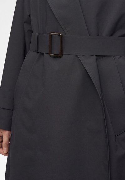 Κοντινό πλάνο σκούρου γκρι παλτού με φαρδιά ζώνη και ορθογώνια αγκράφα, δείχνοντας μέρος του χεριού ενός ατόμου στην αριστερή πλευρά.