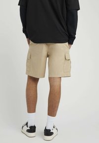 Shorts cargo beiges avec deux poches latérales, fabriqués en tissu coton, présentant une coupe droite et un ourlet. Portés avec des chaussettes blanches et des baskets noires.