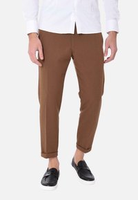 Pantaloni marroni affusolati con una texture liscia; design aderente con risvolti, abbinati a scarpe slip-on nere e una camicia bianca.