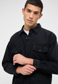 Schwarzes Cordhemd mit Button-Down-Kragen, zwei Brusttaschen und Knopfmanschetten. Es verfügt über eine vertikale gerippte Textur und dezentes Branding.