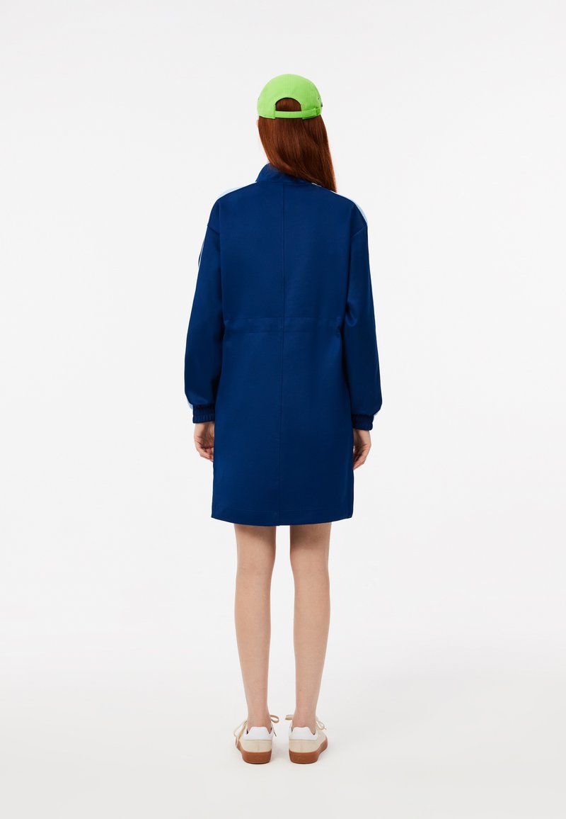 Abrigo corto azul con mangas largas, puños elásticos y corte recto. Lleva puesto un gorro verde y zapatos blancos con suela marrón.