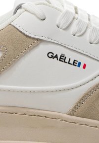 Sneaker in pelle bianca e suede beige con lacci tondi, caratterizzata da un logo con la scritta "GAËLLE" e bandiere francesi in blu e rosso.